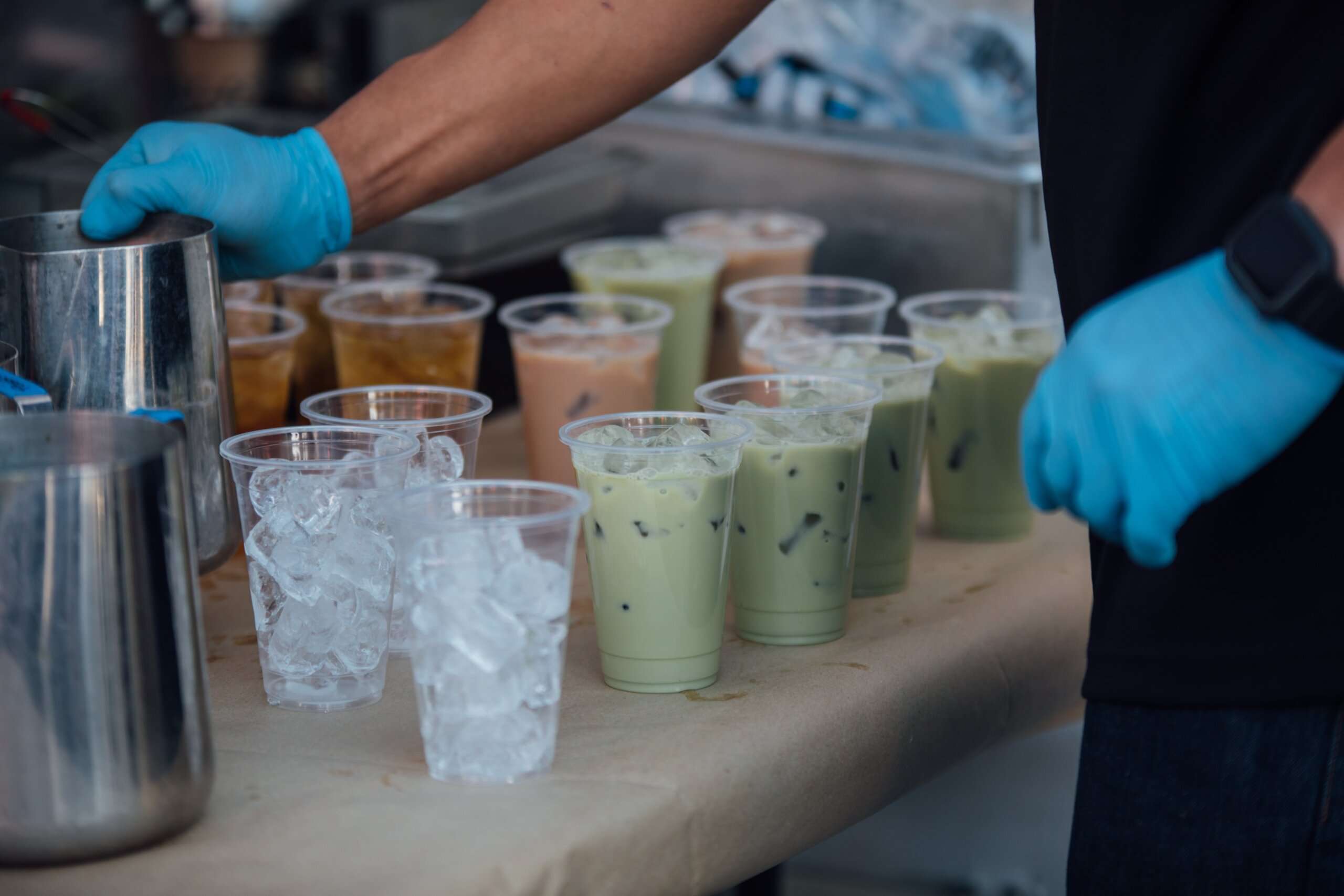an arm with blue plastic gloves reaching out over a bunch of clear plastic cups with ice, green milk tea and brown milk tea in it