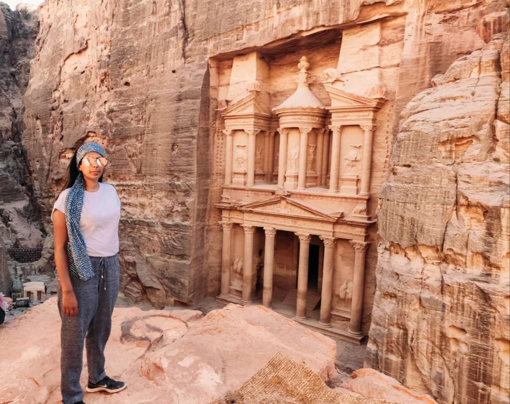 Woman wearing a blue scarf, white shirt and blue pants, standing in front of ancient ruins built into a mountain in the desert