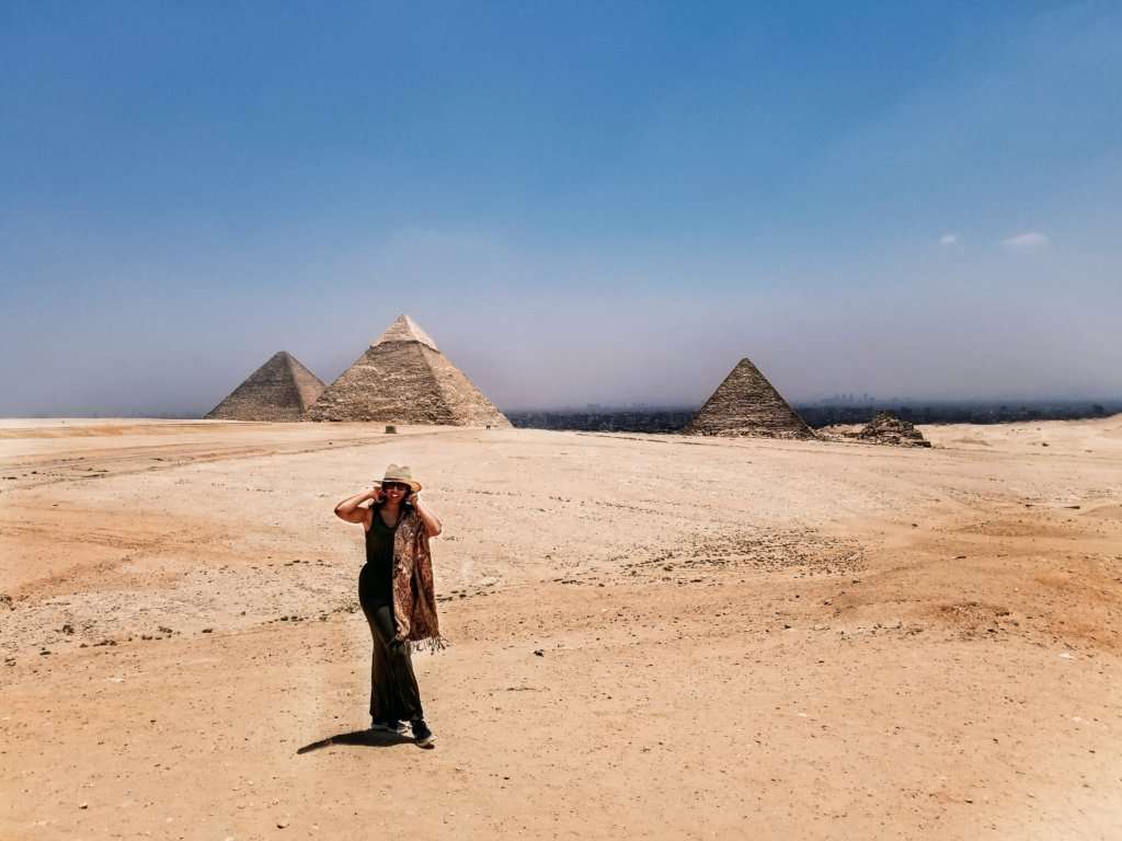Woman standing with hands near her hat standing in front of three stone pyramids in a desert