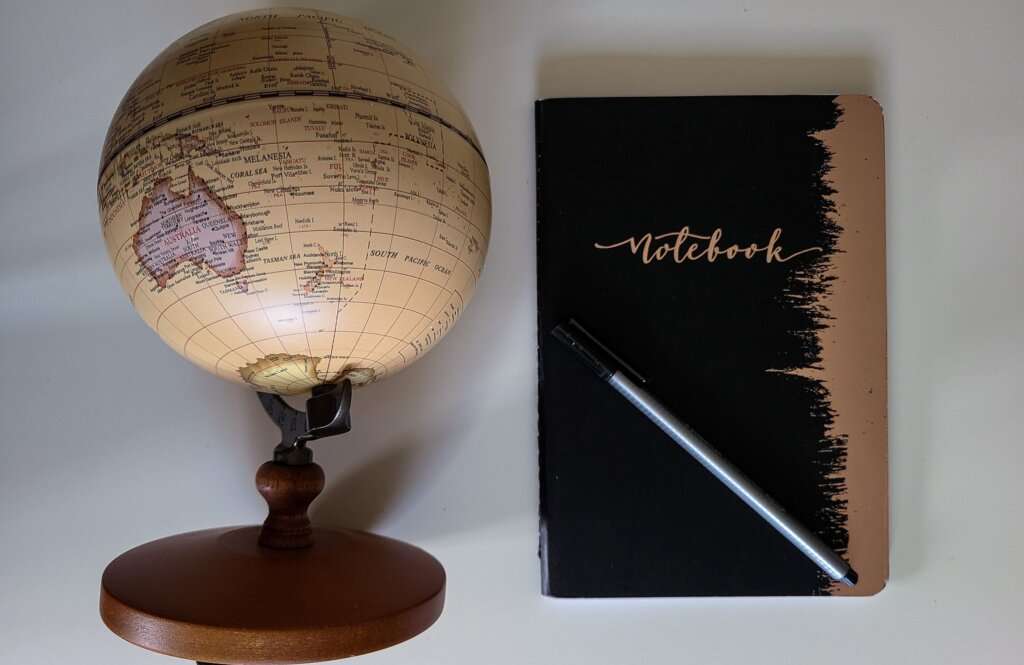 a brown globe next to a notebook that is black with a rose gold border and a pen on top of the notebook