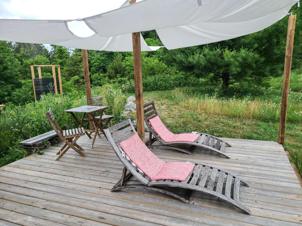 a deck with two lawn chairs with pink cushions and a white tent type cover above the chairs in a green outdoor area