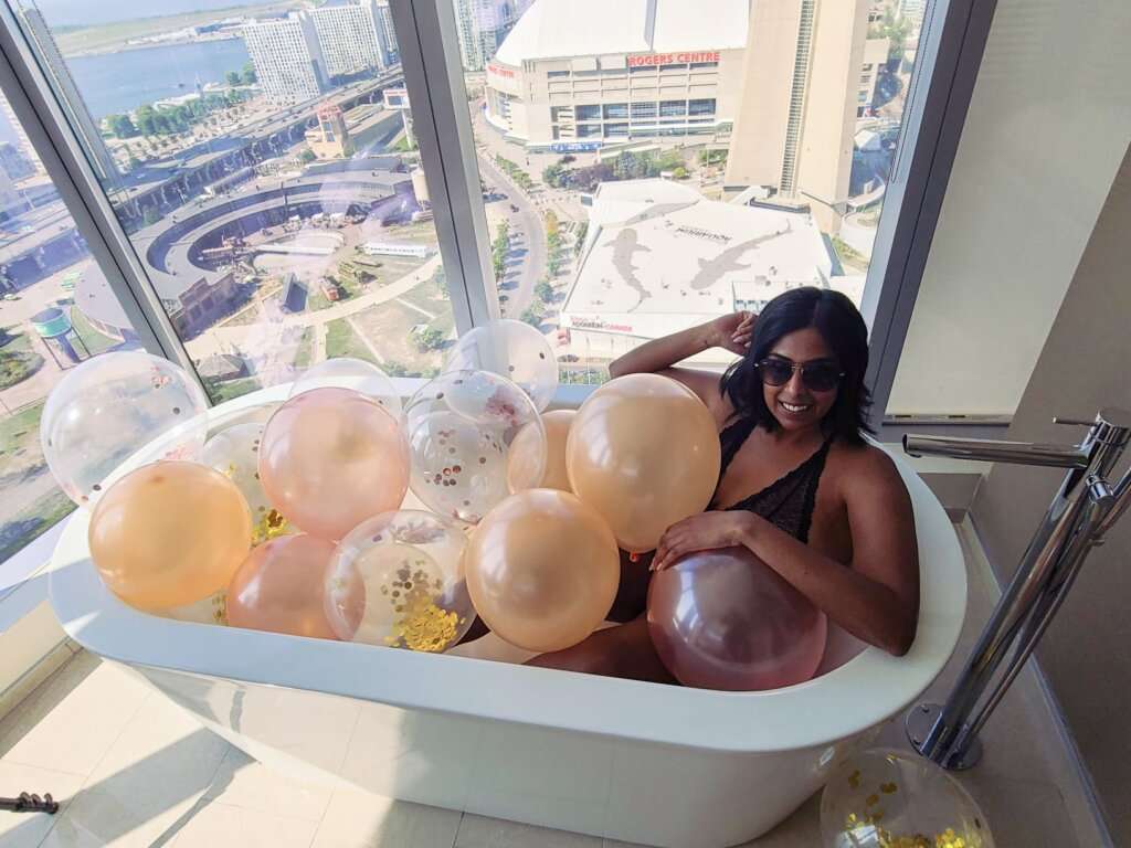 a woman with sunglasses sitting in a bathtub filled with balloons next to a window overlooking a city