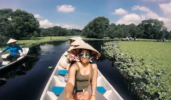 Woman taking a selfie, wearing a bamboo cone hat in a canoe surrounded by green shrubs in the water, with other canoes following.
