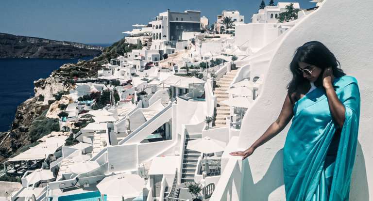 Woman standing with hand in hair wearing a truqiouse blue saree with a white background of stone hotels
