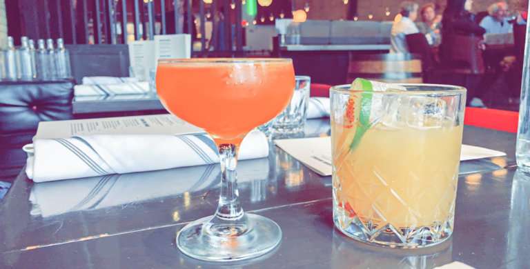 Two drinks on a grey tabletop. One yellow drink and one orange drink.