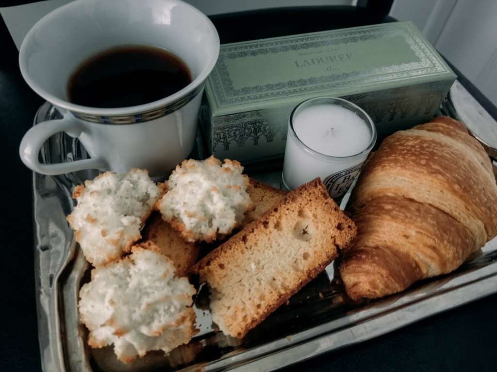 a coffee cup with black coffee, next to a green box, pastries, a croissant and a candle all on a silver carrying tray at home