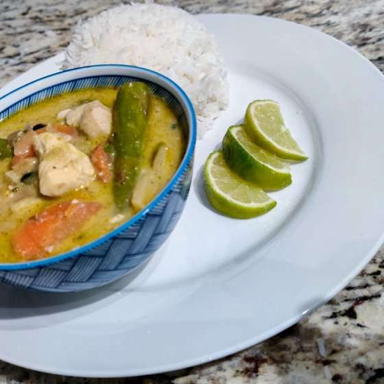 a white plate with whit rush on the side next to three lime wedges and a blue bowl of Thai green curry - on top of a table