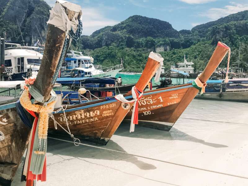 the front of three wooden boats sitting on white sand with blue waters and green hills behind it