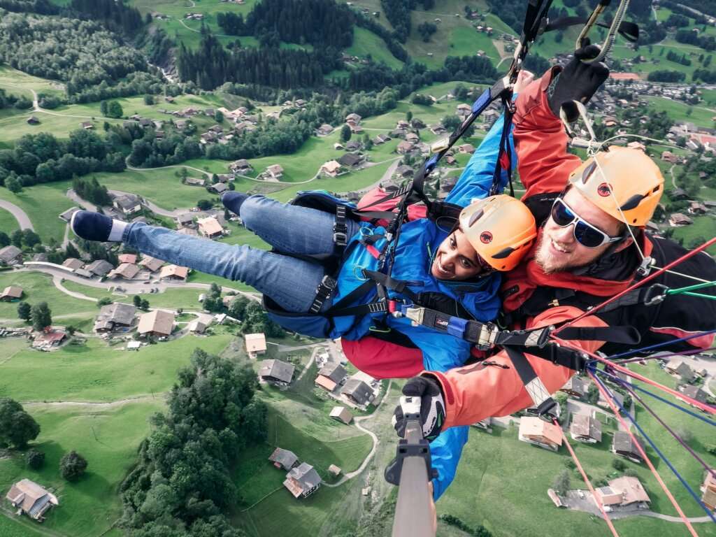 a man in an orange jacket and a woman in a blue jacket in the sky, above houses and trees, paragliding in Switzerland 