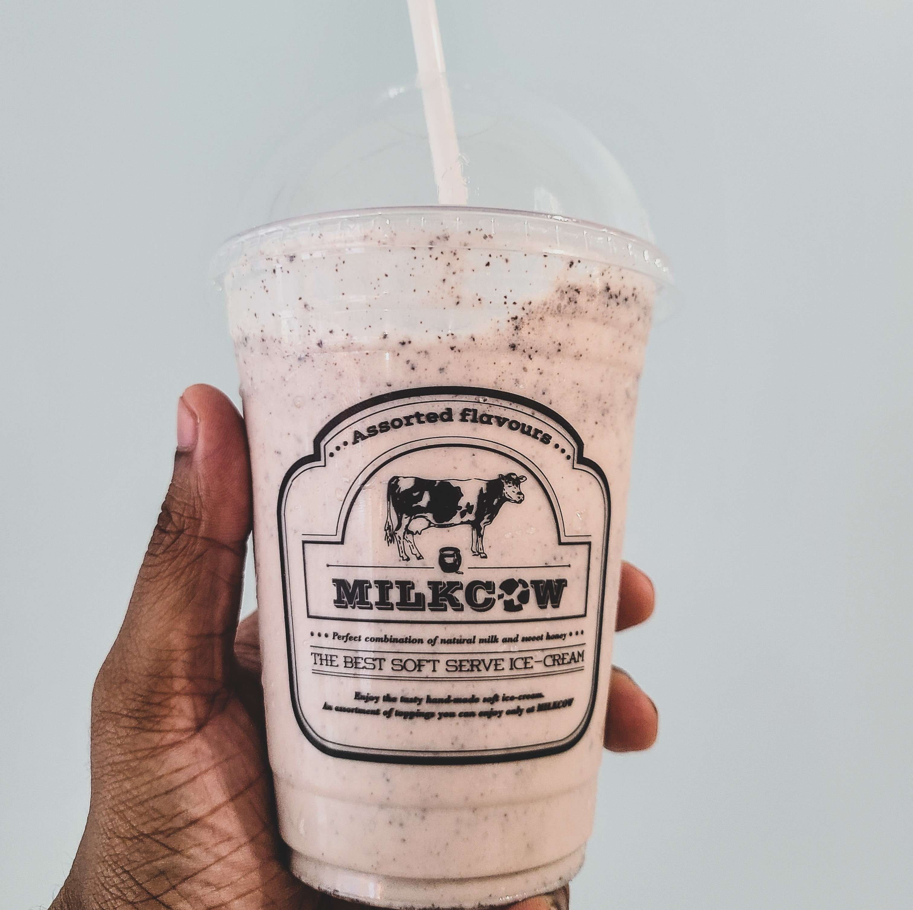 a brown hand holding an oreo milkshake with a white straw from milkcow cafe against a light blue background