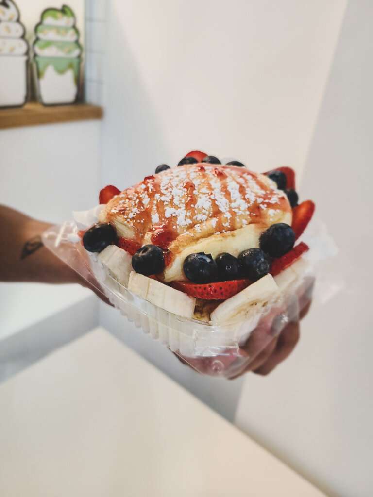 a pancake souffle in a clear plastic container with strawberries, bananas and blueberries - held by a brown hand