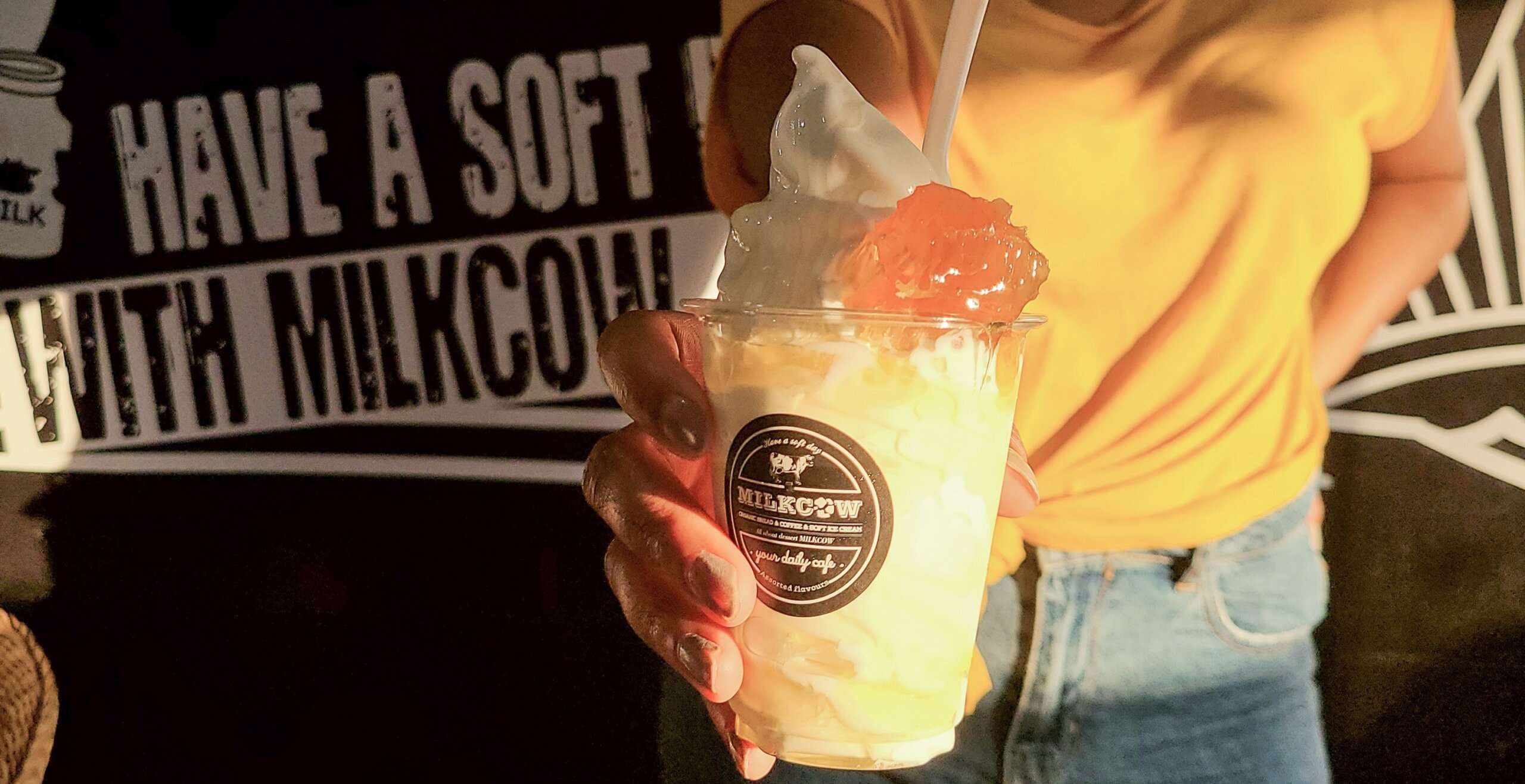 a woman in a yellow top holding a white soft serve ice cream from milkcow cafe in her hand against a black backdrop