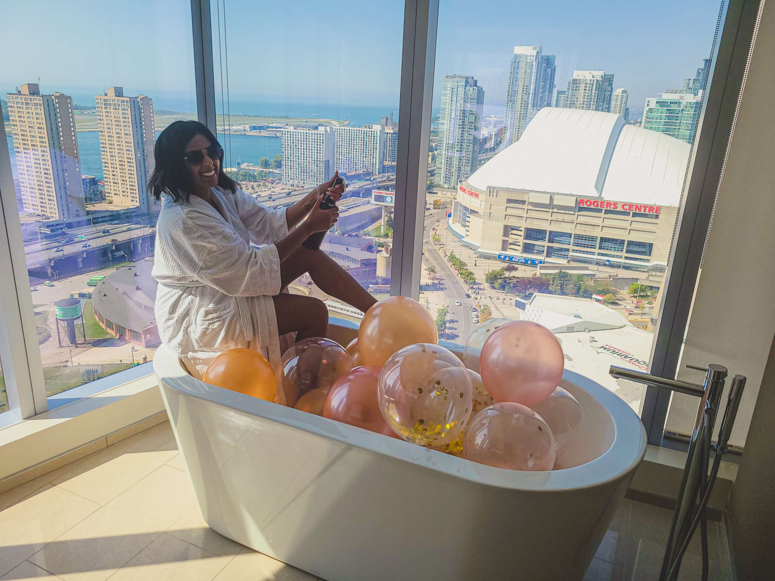 a woman in a white bathrobe sitting on the edge of a bathtub, with large windows overlooking the city with a champagne bottle