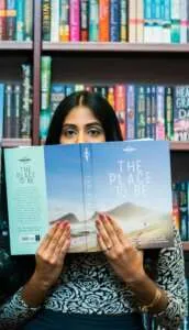 a brown woman standing in front of a bookcase and holding a book while only her eyes peak through the top of the book.