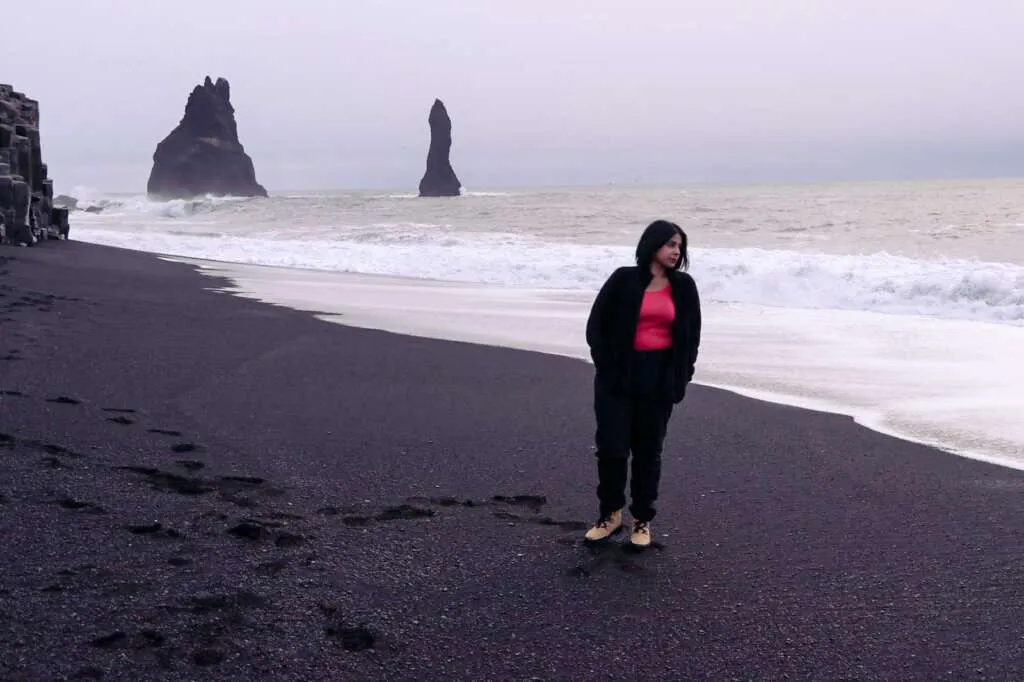 a woman wearing black and pink standing on black sand while looking out at the waters crashing against the ocean.