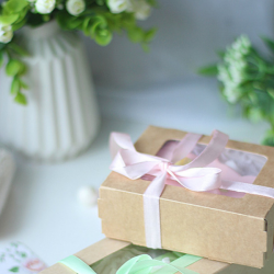 white vase with green leaves and white flower buds and two gifts on a white table wrapped in brown with pink and green ribbon for mothers gifts