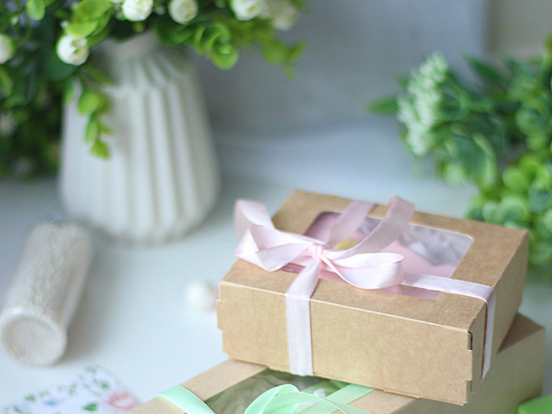 white vase with green leaves and white flower buds and two gifts on a white table wrapped in brown with pink and green ribbon for mothers gifts