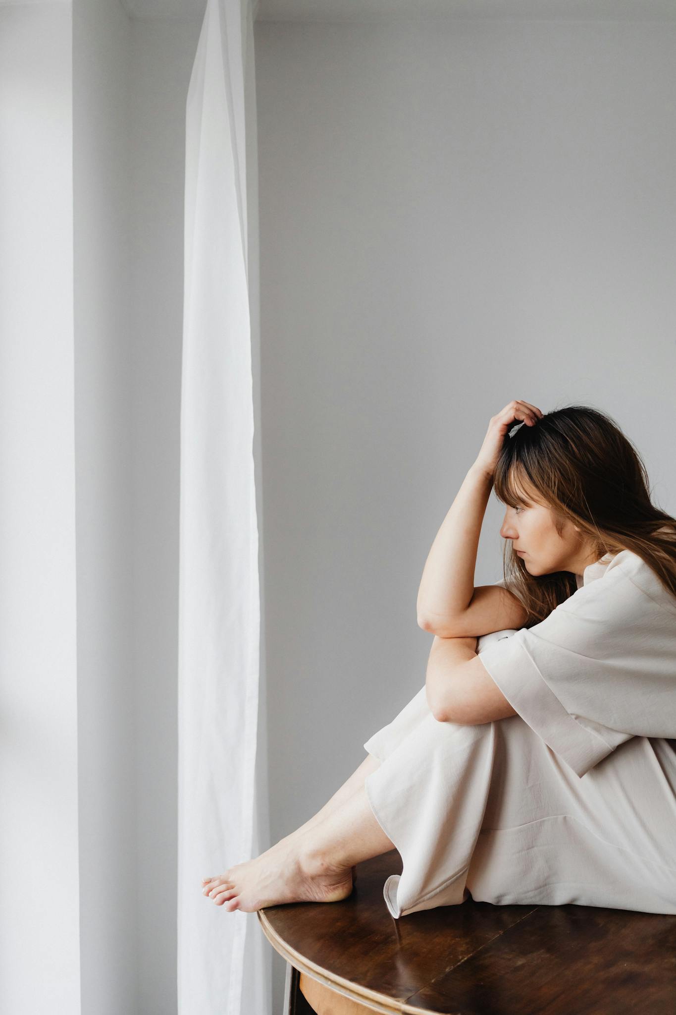 A woman with long hair sits pensively next to a window, creating a serene and introspective scene.